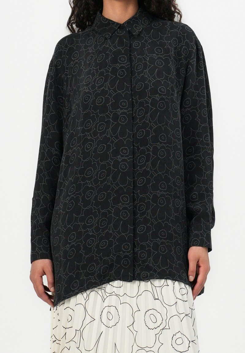 Femme portant une chemise noire à manches longues avec des contours floraux foncés sur une jupe blanche avec de grands imprimés floraux noirs.