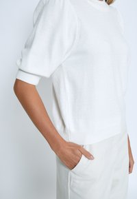 Weißer Strickpullover mit kurzen, gepufften Ärmeln und einem abgerundeten Ausschnitt. Der Stoff wirkt weich und strukturiert, kombiniert mit hellfarbenen Hosen.