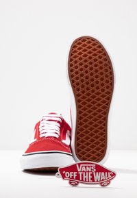Röd kanvas sneaker med vita detaljer, med en gummisula med diamantmönster. Inkluderar en cirkulär "Vans Off The Wall" etikett.