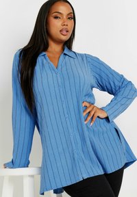 Camicia button-up azzurro chiaro con righe verticali, maniche lunghe, colletto e spacco ai lati per una maggiore libertà di movimento. Finitura in tessuto testurizzato.