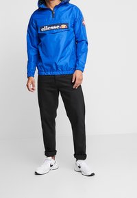 Man som bär en ljusblå ellesse vindjacka, svarta uppvikta jeans och vita Nike-sneakers, stående framför en helt vit bakgrund.