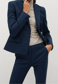 Navy blue suit features a tailored blazer with a single button and flap pockets, paired with slim-fit trousers and a light grey turtleneck.