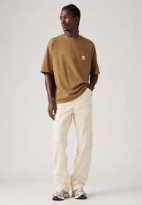 Camiseta de algodón marrón con un bolsillo, de corte holgado, combinada con pantalones color crema y zapatillas plateadas, sobre un fondo neutro.