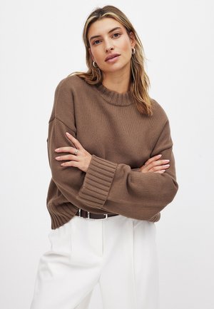 Suéter marrón de punto con un cuello redondo acanalado y mangas extragrandes, combinado con pantalones blancos de tiro alto y un cinturón marrón.