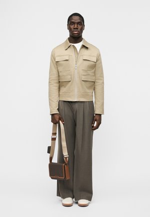 Man draagt een beige jas over een wit shirt, grijze broek met wijde pijpen, witte schoenen, en houdt een bruine crossbody tas vast aan het bandje.