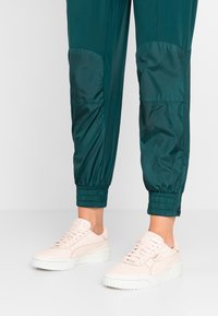 Baskets en daim rose clair avec un design perforé, semelle en caoutchouc blanche et marquage subtil. Portées avec un pantalon de jogging bleu sarcelle à ourlets élastiques.