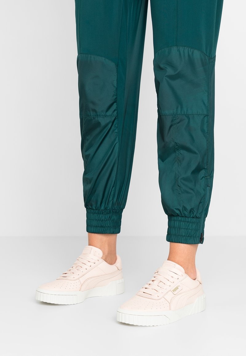 Baskets en daim rose clair avec un design perforé, semelle en caoutchouc blanche et marquage subtil. Portées avec un pantalon de jogging bleu sarcelle à ourlets élastiques.