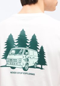 Γραφικό βαν με ψηλά πεύκα και ένα vintage βαν που απεικονίζει το λογότυπο The North Face και το κείμενο «Ποτέ μην σταματάς να εξερευνάς» στην πίσω πλευρά λευκού μπλουζιού.