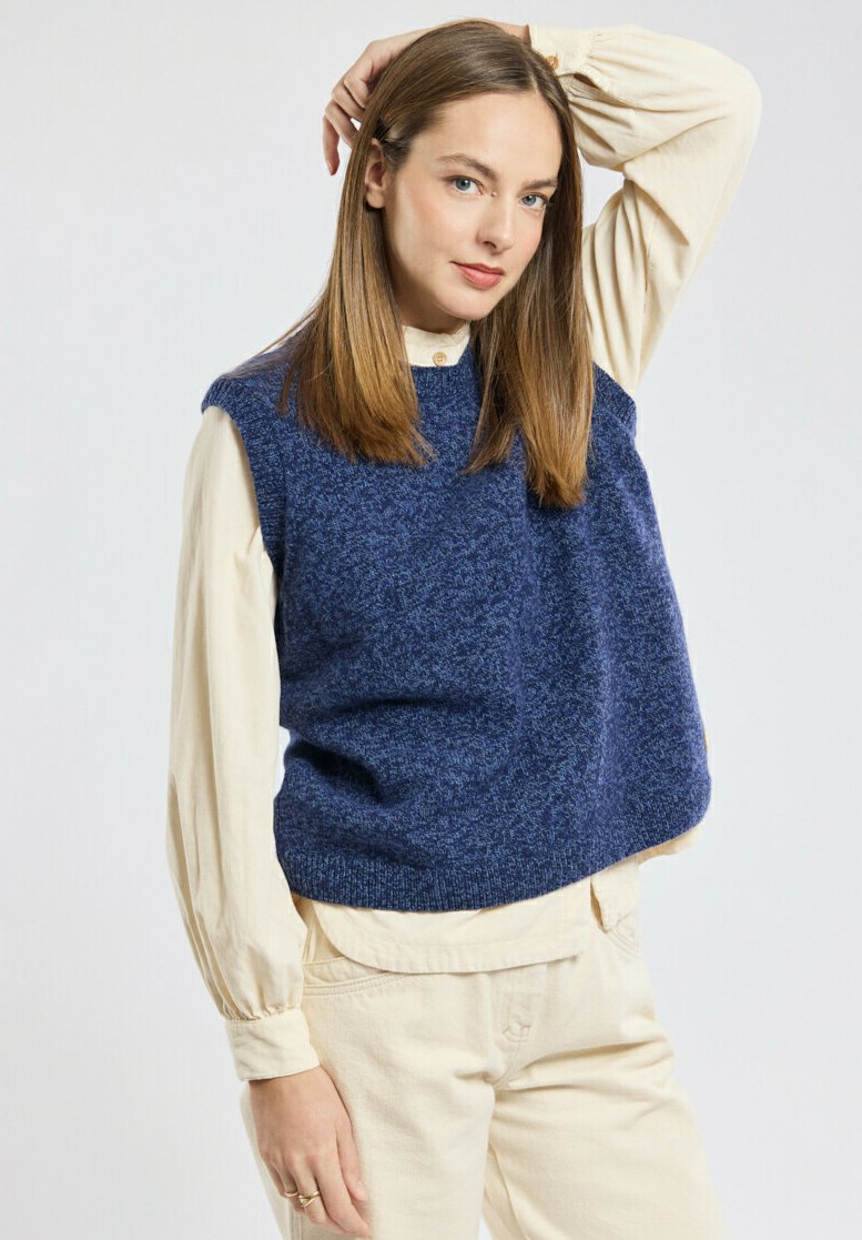 Granatowy, dziany sweter bez rękawów o teksturowanym wykończeniu, noszony na kremowej, długiej koszuli. Jasne spodnie dopełniają outfit.
