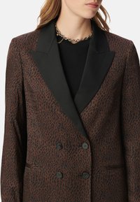 Blazer a stampa leopardo marrone con risvolti in satin nero, bottoni doppi e finitura testurizzata. Design su misura con tasche laterali.
