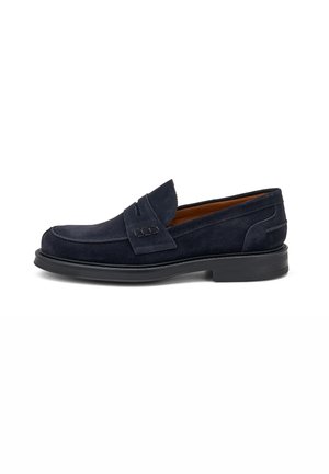 Mocasines de ante azul marino con punta redondeada, costuras decorativas y una suela baja de goma negra. Diseño clásico sin cordones con acentos sutiles.