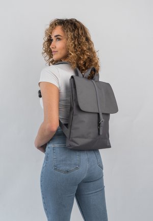 Gray backpack made of synthetic fabric, featuring a flap closure, adjustable straps, and minimalistic design. Worn on a person's back.