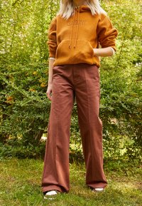 Orange fleece-hoodie med frontficka och dragsko, kombinerad med höga, vida bruna byxor, mot en grön bakgrund.