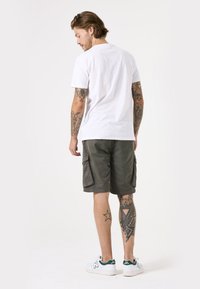 Camiseta de algodón blanca, pantalones cortos cargo verdes con bolsillos laterales y zapatillas blancas con detalles verdes. Tatuajes detallados visibles en las piernas.