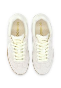 Zapatillas de ante blancas con cordones crema, un logo dorado en la lengüeta y una suela de goma texturizada. Presenta un diseño suave y minimalista.