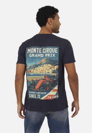 Uomo con maglietta blu navy con grafica in stile vintage di un’auto da corsa rossa su una pista da corsa costiera e testo "Monte Cirque Grand Prix".