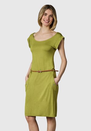 Kvinne smilende iført en kortermet grønn kjole med sidelommer og tynn brun belte, stående med hendene i lommene.