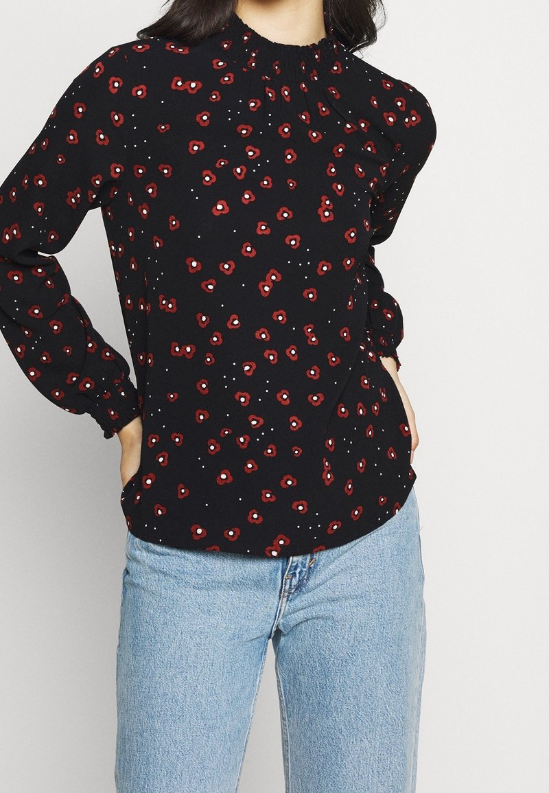 Blusa nera con motivo floreale rosso e dettagli bianchi, caratterizzata da un colletto arricciato e maniche lunghe con polsi elasticizzati. Indossata con jeans blu.