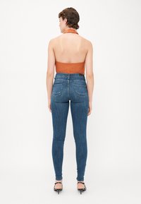 Kék, magas derekú skinny farmernadrág sima denim textúrával, két hátsó zsebbel és enyhe kopással, egy narancssárga pántos top mellett.
