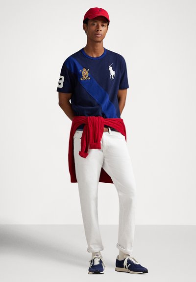 Námořnické polo triko s diagonálním modrým pruhem, červenými akcenty a vyšitým logem, v kombinaci s bílými džínami a červeným svetrem uvázaným kolem pasu.
