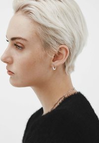 Pendiente de botón geométrico plateado en forma de V. Cabello corto y de color claro; lleva un suéter negro texturizado con un collar de cadena de metal simple.