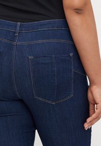 Jeans de un azul oscuro con un diseño clásico de cinco bolsillos y detalles de costura sutiles. La tela tiene una textura de denim suave.
