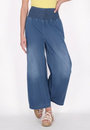 Denim-Hosen mit weitem Bein und smocktem Bund in Mittelblau. Der Stoff hat eine weiche Textur. Kombiniert mit Riemchensandalen und einem gelben Oberteil.