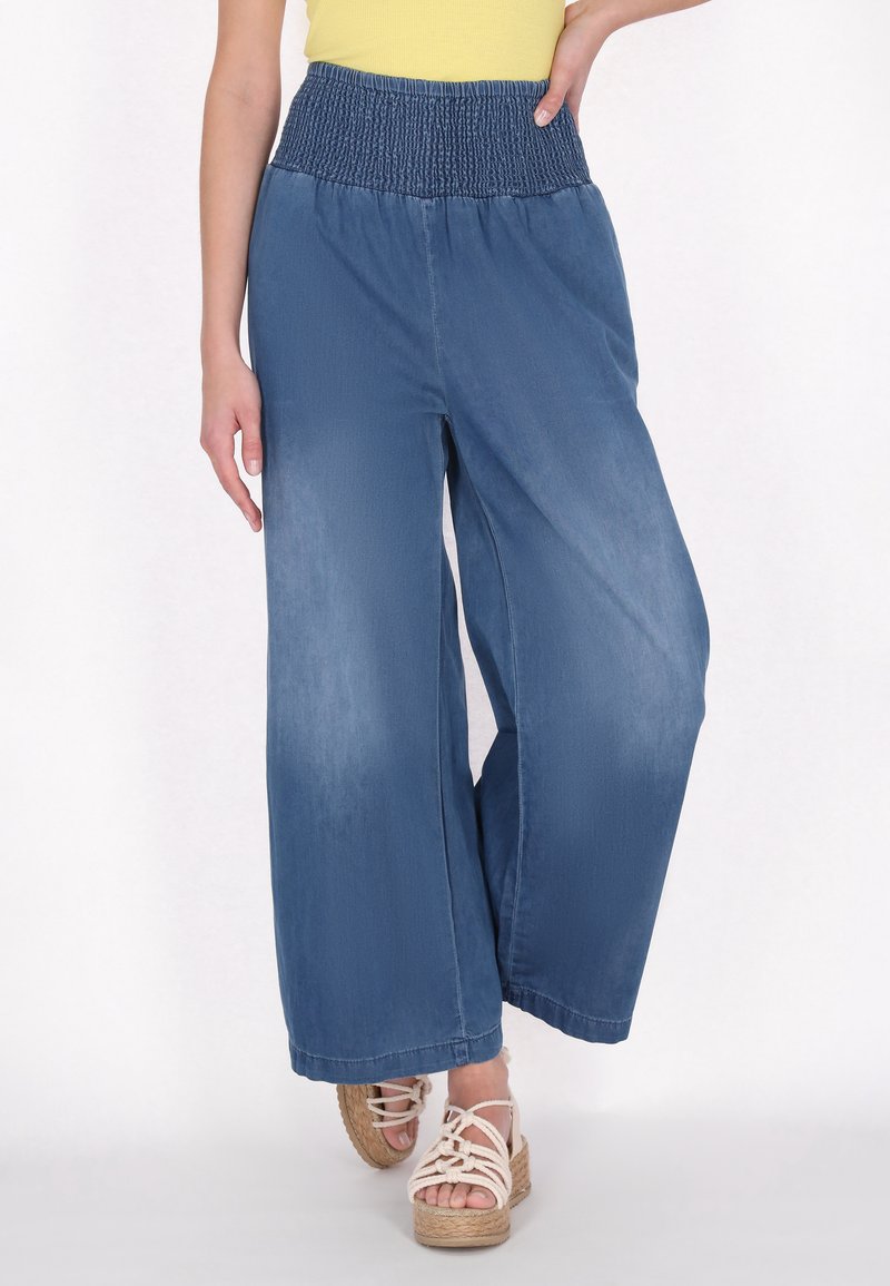 Középkék, bő szárú farmernadrág, smockolt derékkal. Az anyag puha tapintású. Színes szandálokkal és sárga toppal párosítva.