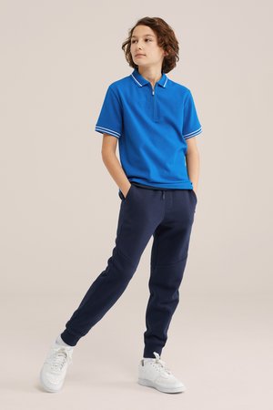 Bambino con capelli ricci in piedi con le mani nelle tasche, indossa una polo blu a maniche corte, pantaloni da jogging blu navy e scarpe da ginnastica bianche.