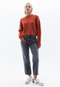 Cropped, oranje gebreide trui met een geribbelde textuur, gecombineerd met hoge, grijze spijkerbroek en witte sneakers met zwarte accenten.