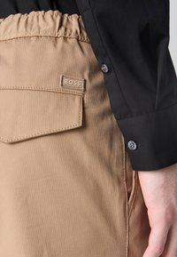 Béžové kalhoty z plisované látky s elastickým pasem a našitým kapsou. Černá košile s knoflíkovými detaily na manžetě.