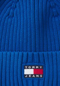 Blå stickad mössa med ribbad textur, prydd med en rektangulär logotyp som visar "TOMMY JEANS" och en färgskala i röd, vit och blå.