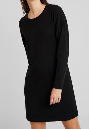 Woman wearing a black long-sleeve knee-length sweater dress against a plain light gray background.