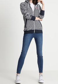 Marineblauw-wit gestreepte hoodie met ritssluiting en capuchon, geribbelde manchetten en zoom, gecombineerd met donkerblauwe skinny jeans en witte sneakers.