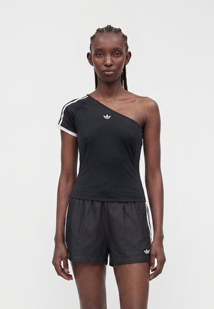 Femme aux cheveux tressés portant un haut Adidas noir à une épaule et un short Adidas noir avec des bandes blanches sur un fond uni.