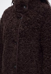 Veste sherpa marron texturée avec des boutons noirs et des poches latérales, portée par une personne aux longs cheveux foncés.