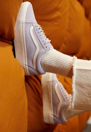 Baskets en suède lavande avec une bande blanche ondulée portées avec des chaussettes blanches côtelées et un pantalon blanc aux bords effilochés sur un fond orange.