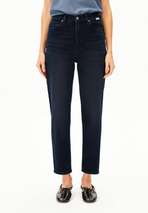Person trägt dunkelblaue High-Waist-Jeans mit geradem Bein und schwarzen Schnürschuhen und steht vor einem einfachen weißen Hintergrund.