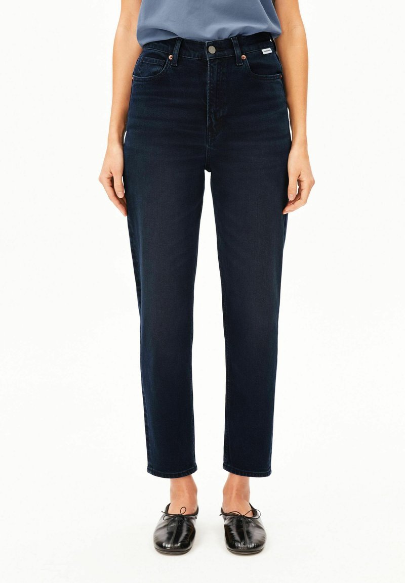 Person trägt dunkelblaue High-Waist-Jeans mit geradem Bein und schwarzen Schnürschuhen und steht vor einem einfachen weißen Hintergrund.