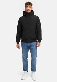 Zwarte gewatteerde jas met een hoge kraag, voorzakken en een verstelbare capuchon, gedragen over lichtblauwe jeans en witte sneakers.