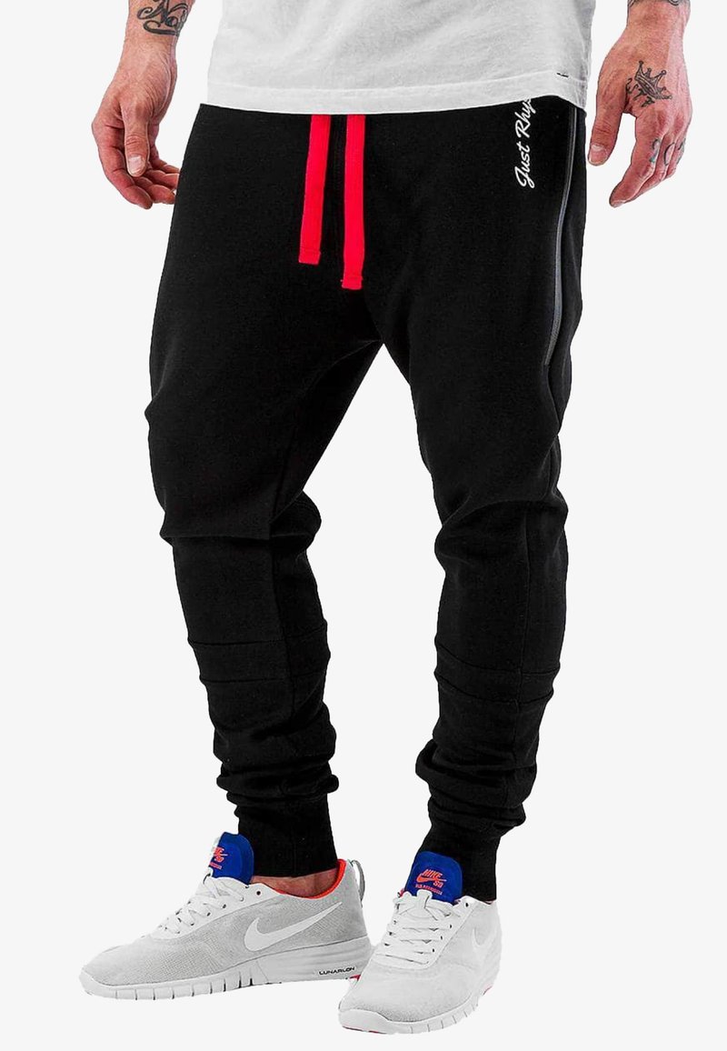 Black jogger pants with red drawstring, ribbed cuffs, and side pockets. Features white brand text on the left thigh. Worn with gray sneakers.