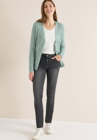 Uśmiechnięta kobieta w jasnoniebieskim dzianinowym kardiganie, białej koszulce z dekoltem w serek, ciemnoszarych dżinsach i białych tenisówkach, stoi z jedną ręką w kieszeni.