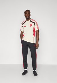 Sportssæt bestående af en cremegul og rød kortærmet skjorte med logo og røde detaljer, parret med mørkegrå bukser med røde striber.