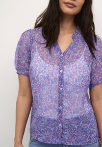 Blusa trasparente viola con motivo floreale, maniche a palloncino corte, chiusura a bottoni e scollatura a V. Tessuto morbido con tessuto leggero e fluido.