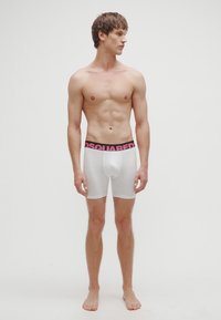 Bóxers blancos con una cintura elástica negra y rosa con la marca, que ofrecen un ajuste ceñido y una textura suave. El modelo está descalzo y posa frente a un fondo liso.