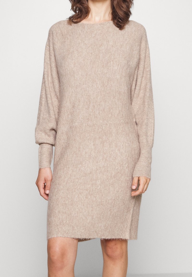 Femme portant une robe-pull beige à manches longues en maille, debout devant un fond clair uni.