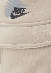 Beige Nike joggers van rekbaar materiaal, met een opvallend zwart logo en gestikte zakken, die de gladde textuur en het moderne ontwerp benadrukken.