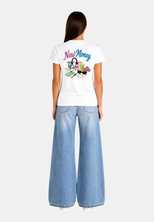 Donna che indossa una t-shirt bianca con testo colorato "New Money" e grafica di oggetti di uso quotidiano, abbinata a jeans a gamba larga azzurri.