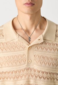 Polo beje de malha com um padrão de renda, apresentando um colarinho e três botões. Colar prateado com um encantamento decorativo.