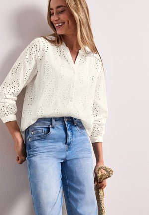 Femme souriante portant une blouse blanche à œillets avec boutons et un jean bleu clair, tenant un sac beige tissé dans sa main droite.